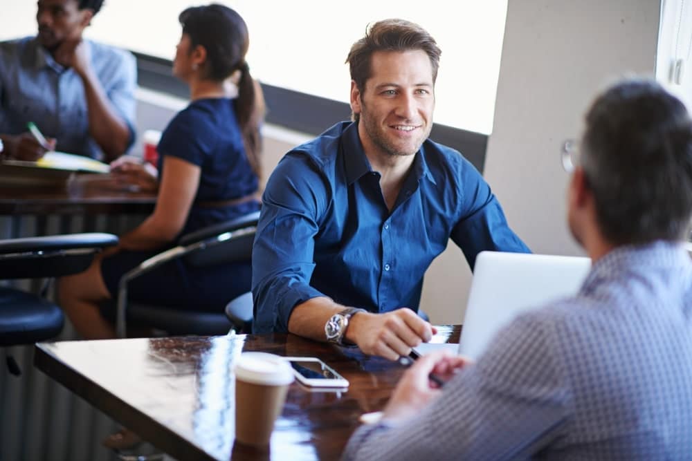 Two businessmen at coffee table in crowded` cafe with coffee cup, smartphone and laptop discuss recruitment solutions
