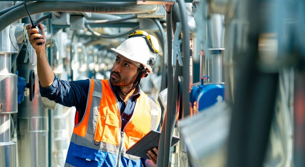 Construction Inspector in Orange Safety Vest Holds Radio and Tablet and Inspects Piping