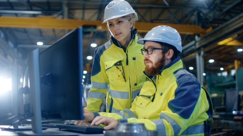Two plant workers in hazard vests and hard hats working at laptop reviewing production manager jobs and responsibilities