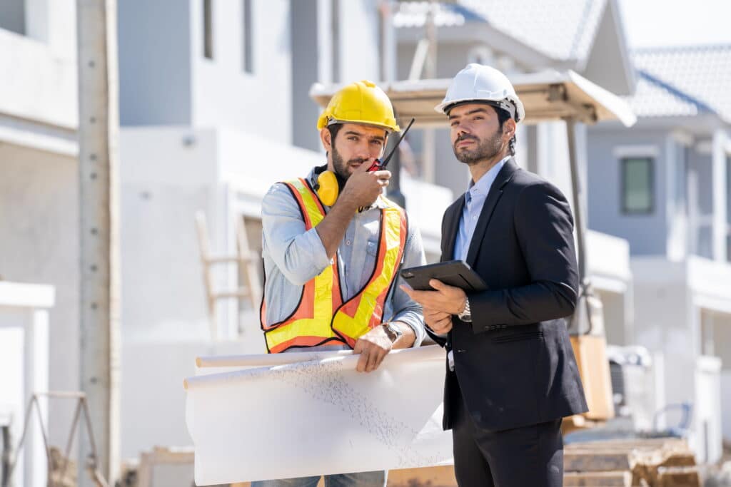 A construction foreman in a safety vest and a project manager in a suit discussing project blueprints on a job site, illustrating how to find headhunters for the construction industry to recruit top executive talent.