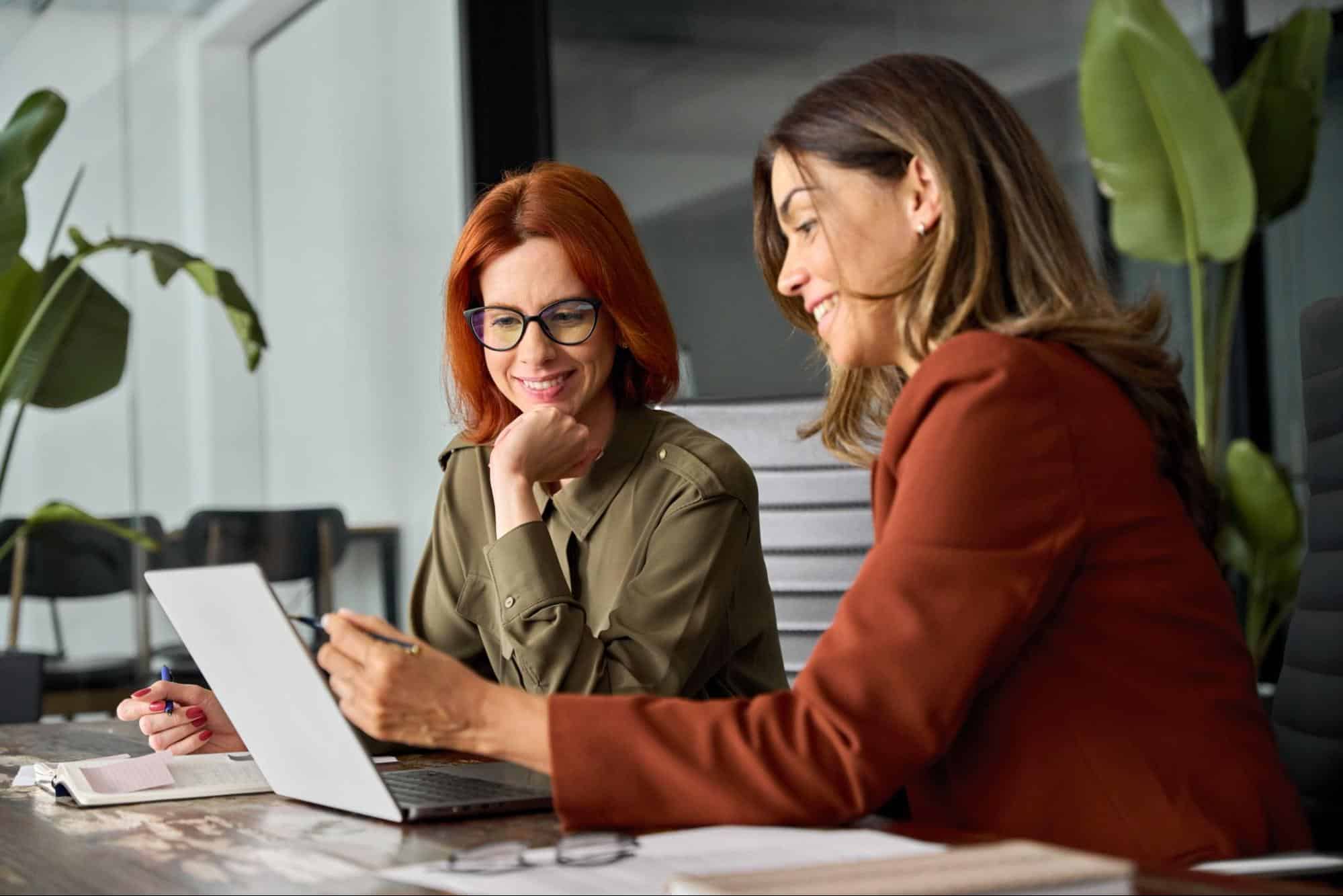 Two professional women collaborating over a laptop in a bright office, implementing LinkedIn profile tips to optimize their digital presence for executive recruiters.