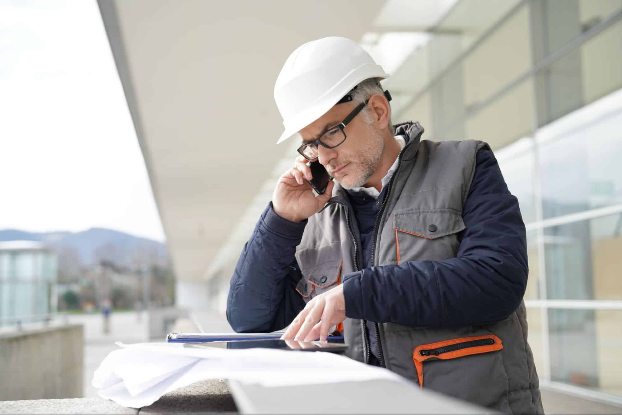 Construction Project Manager in White Hard Hat Reviewing Documents Outdoors in Front of Windows