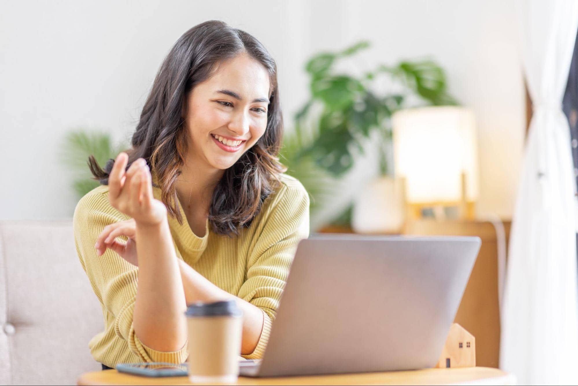 A happy young woman sits at a bright desk with a coffee cup, gesturing and looking at her laptop screen as she uses AI resume builders to craft her job application.