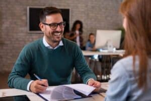 A smiling male interviewer in a green sweater sits at an office desk, holding a pen and reviewing AI-generated resumes on a clipboard across from a female job candidate.