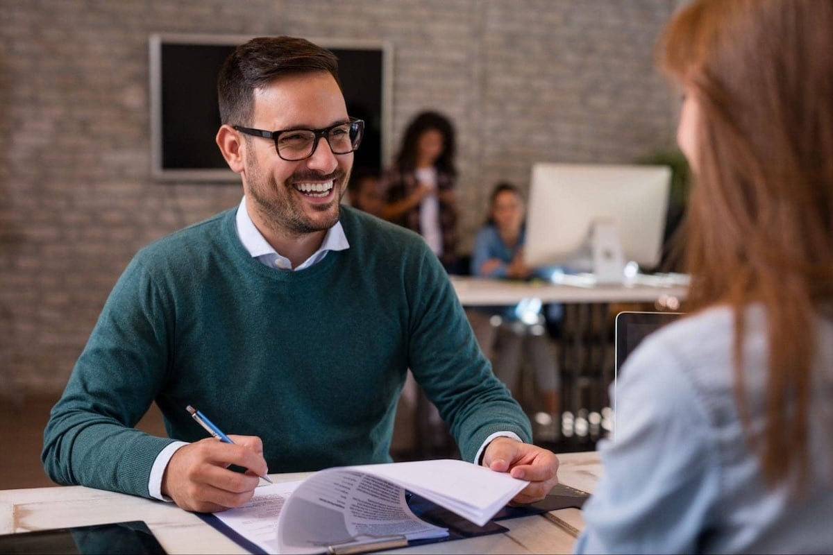 A smiling male interviewer in a green sweater sits at an office desk, holding a pen and reviewing AI-generated resumes on a clipboard across from a female job candidate.