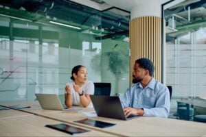 Two corporate staffing consultants discussing talent placement over laptops in a modern glass office, highlighting the collaborative process used by professional HVAC recruitment agencies.