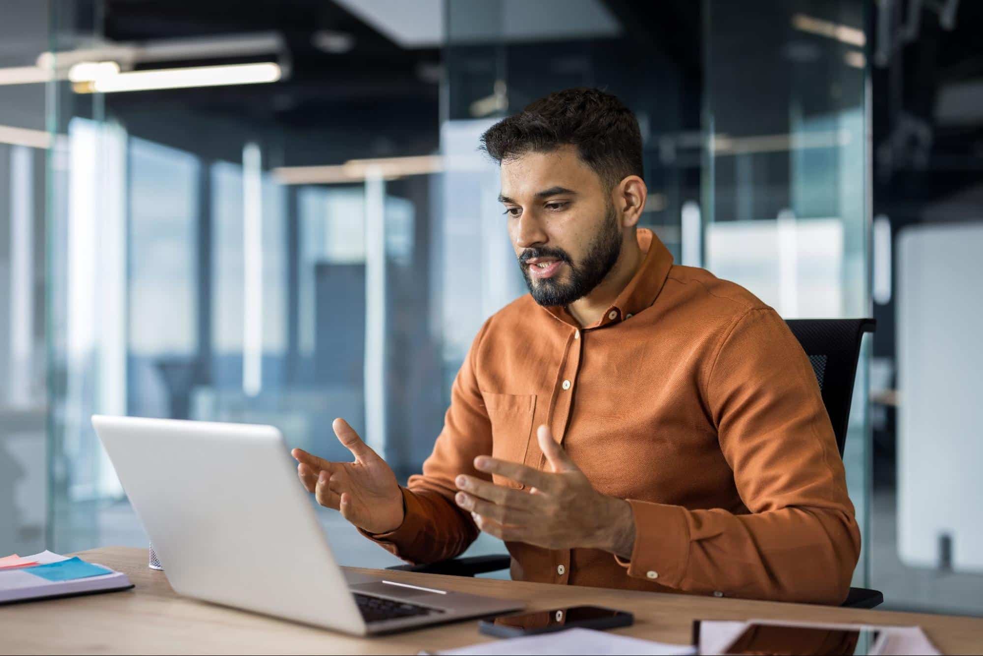A male professional in an orange shirt gesturing during a video call on his laptop in a modern corporate office, illustrating a successful candidate interview for an HVAC executive search.