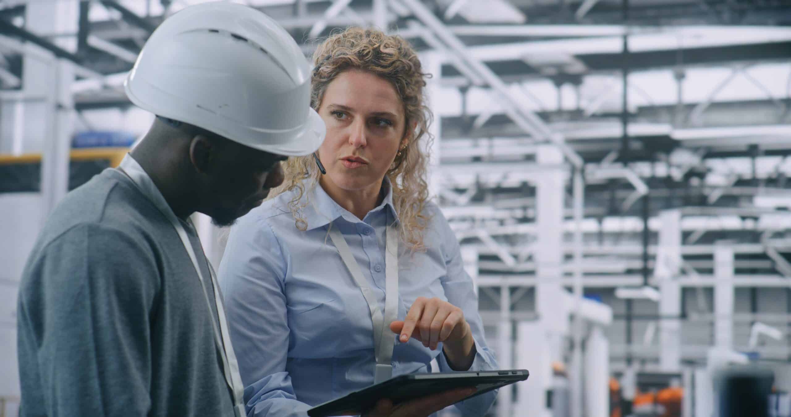 A female manager pointing to a tablet while discussing operational plans with a male technician wearing a white hard hat on a manufacturing plant floor.
