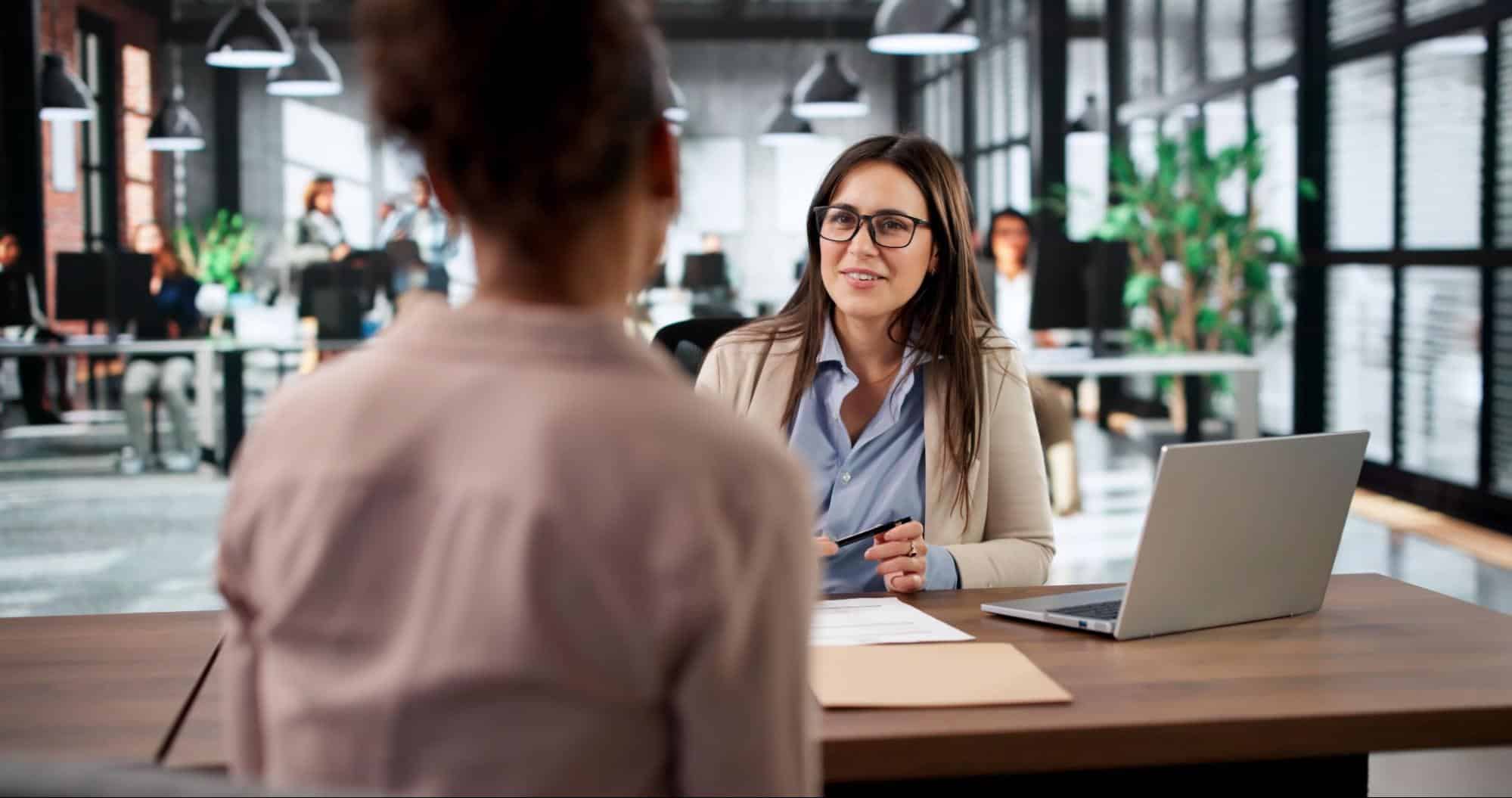 A hiring manager with glasses smiles warmly while conducting a talent acquisition interview across a desk in a modern open-concept office.