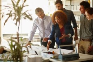 Five real estate executives standing around a conference table discussing building plans in a bright office.